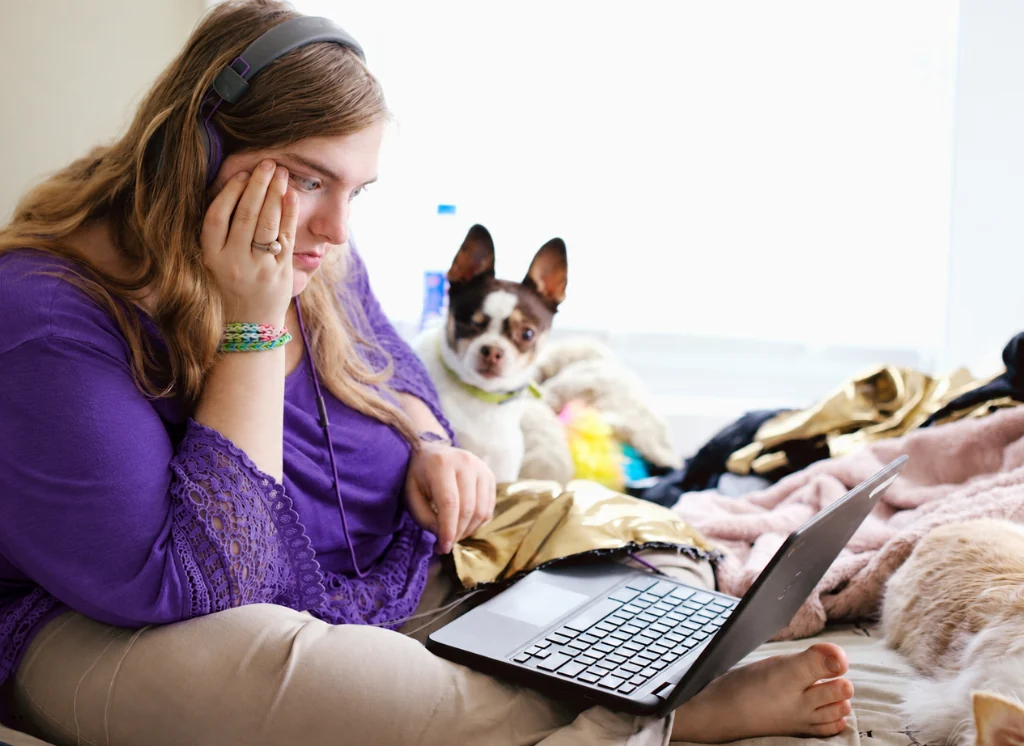 A woman studying on a laptop at home with a small dog beside her